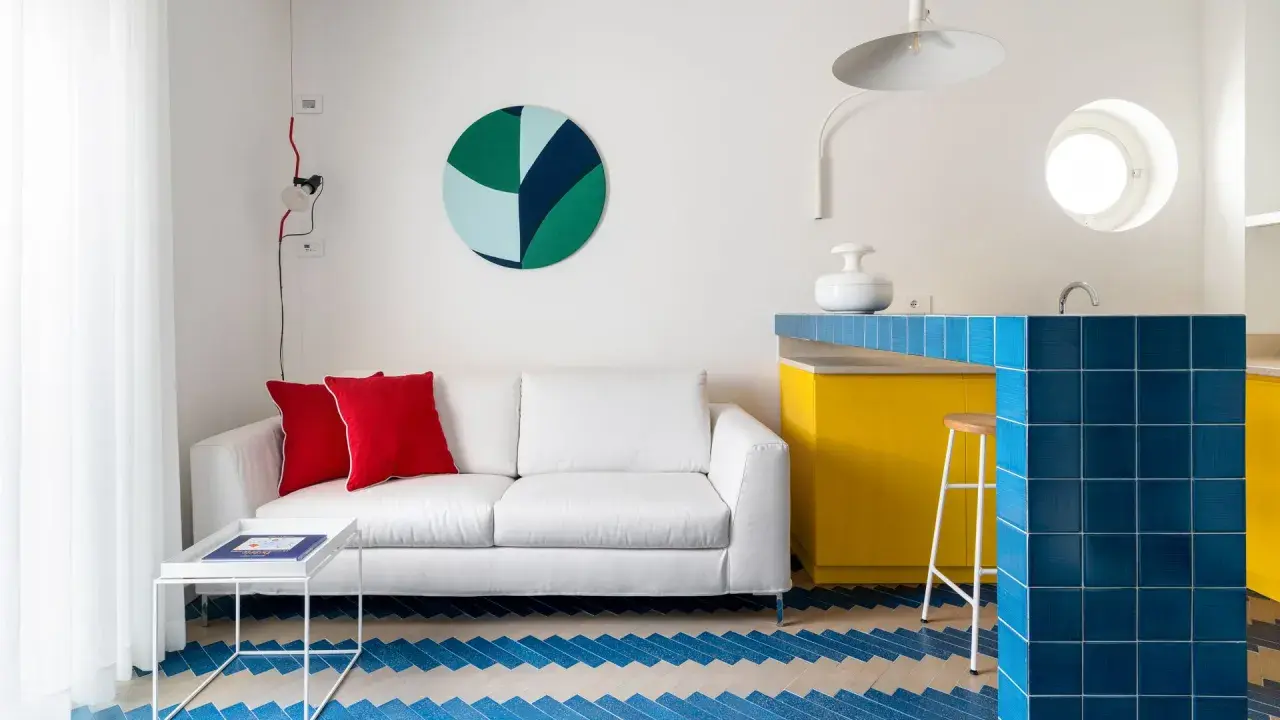 Sofá blanco con cojines rojos, mesa auxiliar blanca y suelo con diseño de espiga en colores claros y azul. Barra de cocina amarilla y azul.