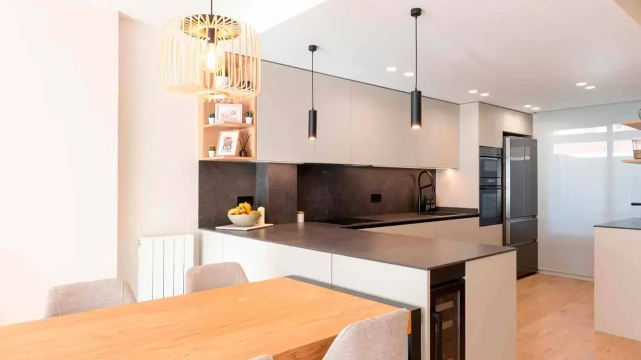 Moderna cocina en L con isla, electrodomésticos integrados, encimera de cuarzo y mesa de madera.