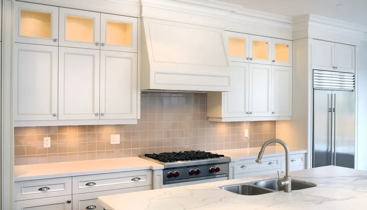 Cocina moderna con campana extractora decorativa sobre estufa. Gabinetes blancos, encimera de mármol y refrigerador de acero inoxidable.