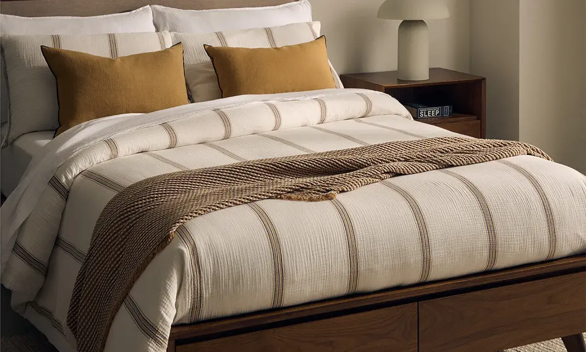Cama con ropa de cama a rayas, cojines mostaza y manta tejida. Las bassols sabanas aportan un toque acogedor.