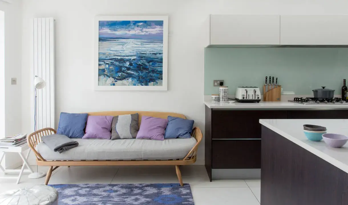 Acogedora cocina con un cómodo sofá de madera y cojines coloridos, junto a una isla y un cuadro de paisaje marino.