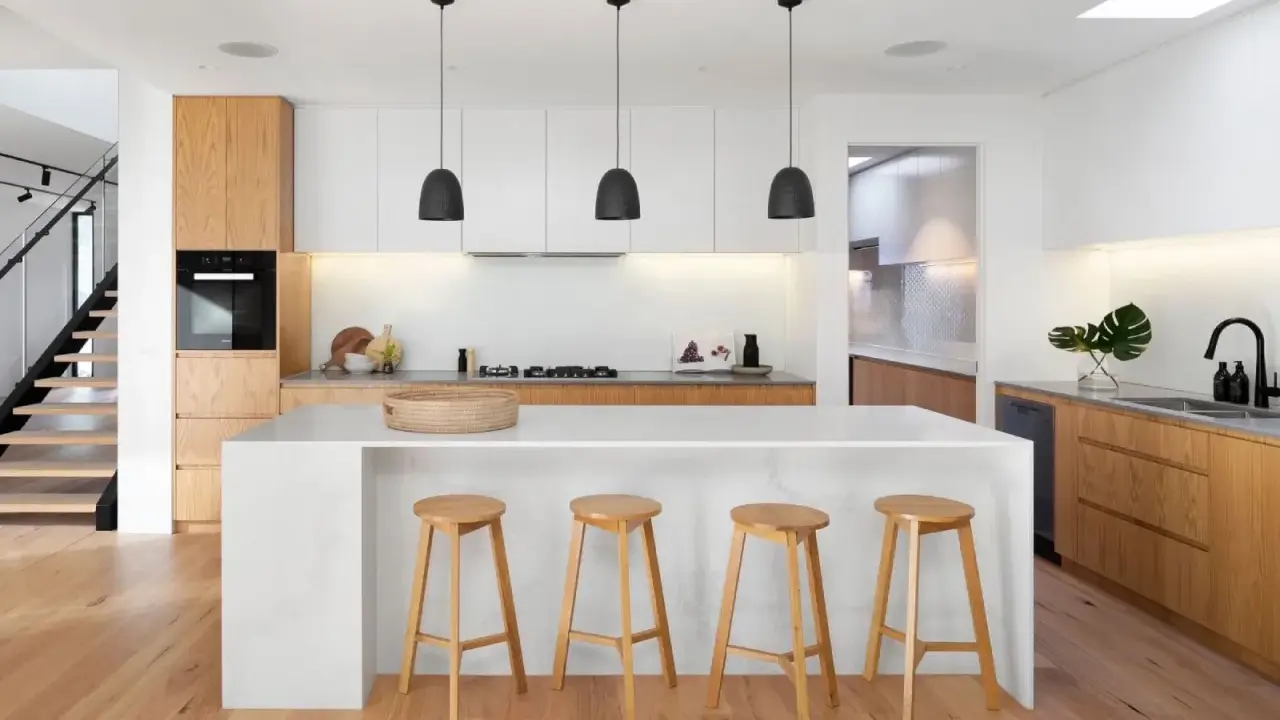 Cocinas modernas con isla blanca, taburetes de madera y gabinetes de roble. Escalera de diseño minimalista al fondo.