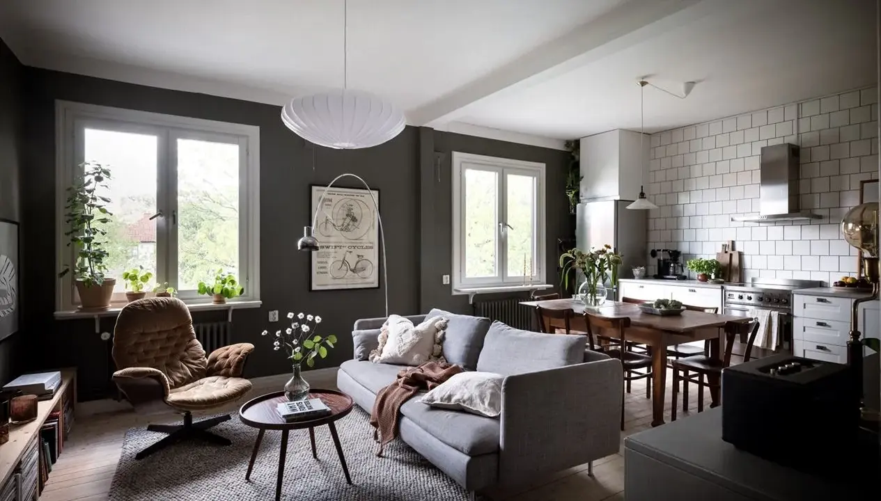 Un salón rectangular amueblado con sofá gris, sillón marrón, mesa de centro y alfombra. Cocina abierta con mesa de comedor.