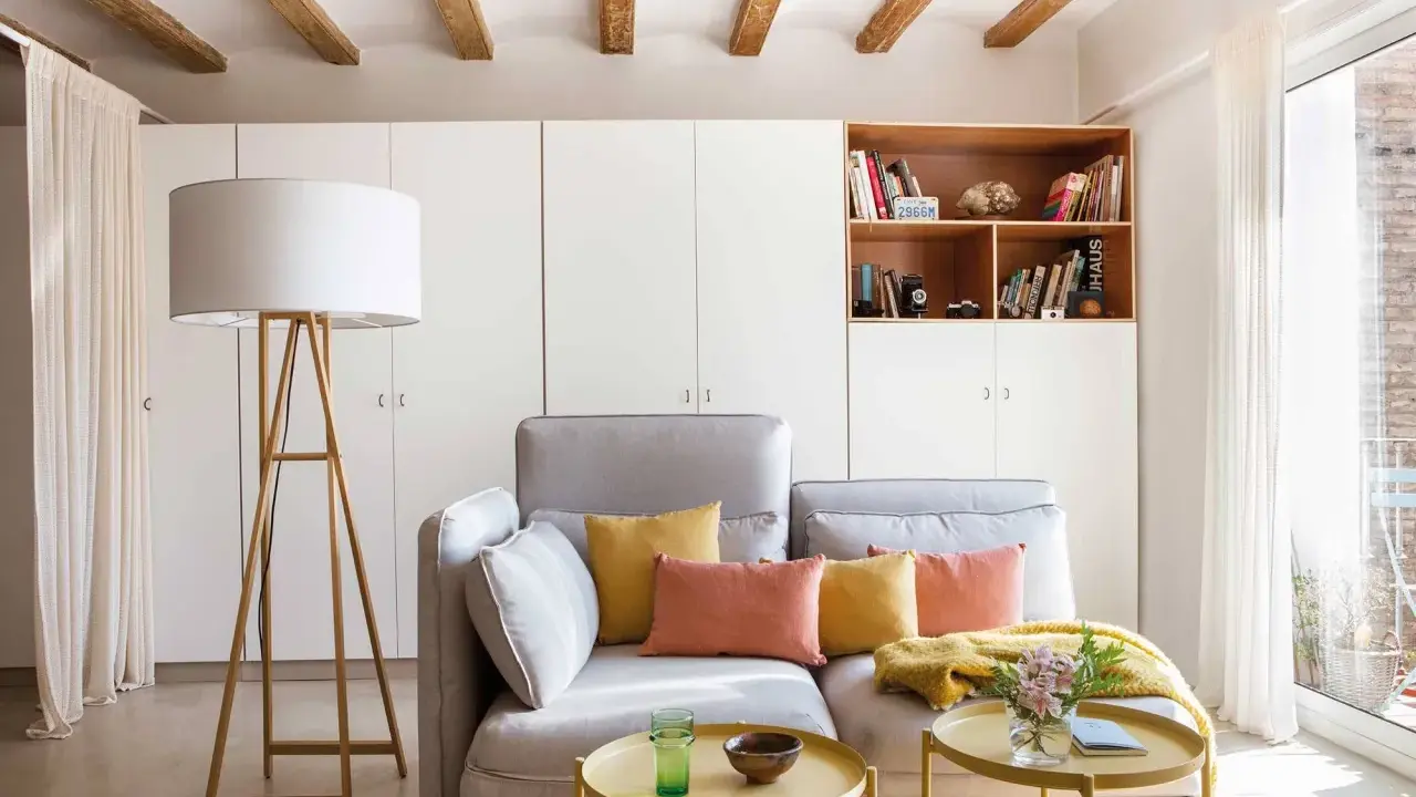 Ideas para decorar piso alquiler low cost: sofá gris, cojines coloridos, mesa auxiliar dorada, lámpara de pie y estantería con libros.