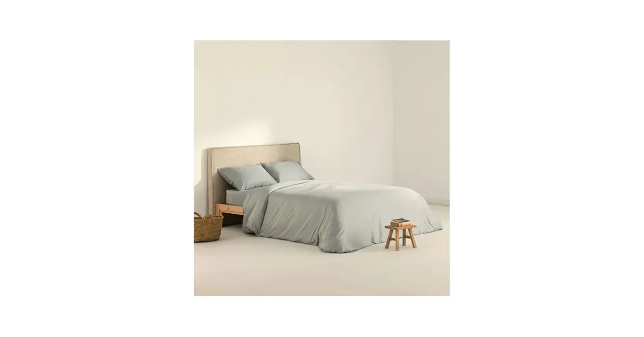 Cama con funda nordica 180x220 en tono gris claro, almohadas a juego y una cesta de mimbre al lado.