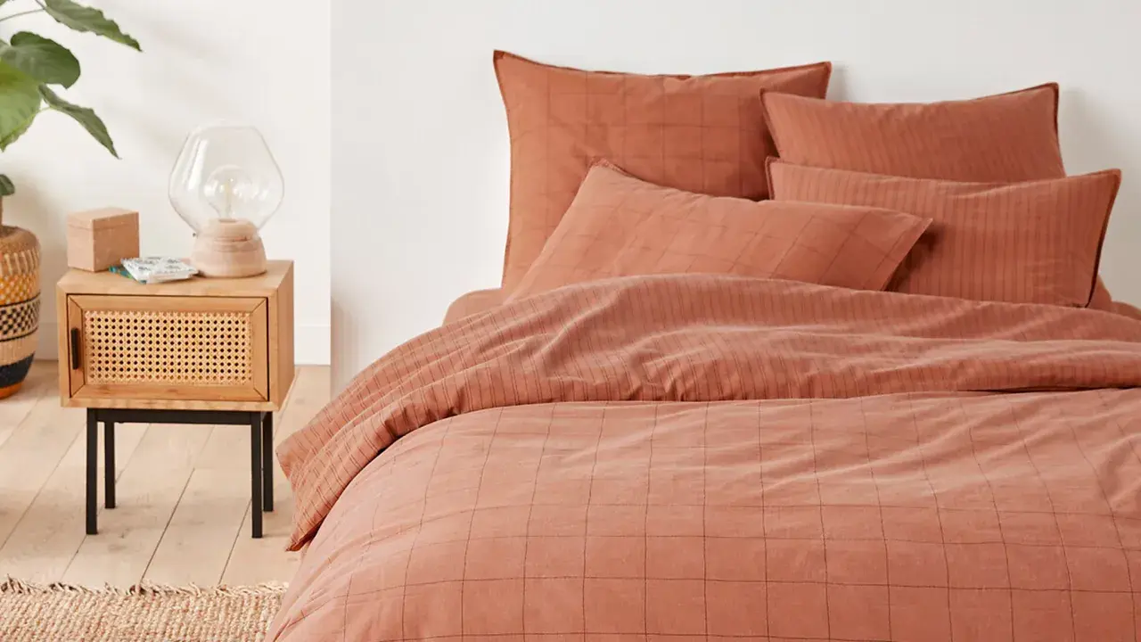 Cama con funda nordica terracota a cuadros y rayas. Mesita de noche de madera con lámpara y planta.