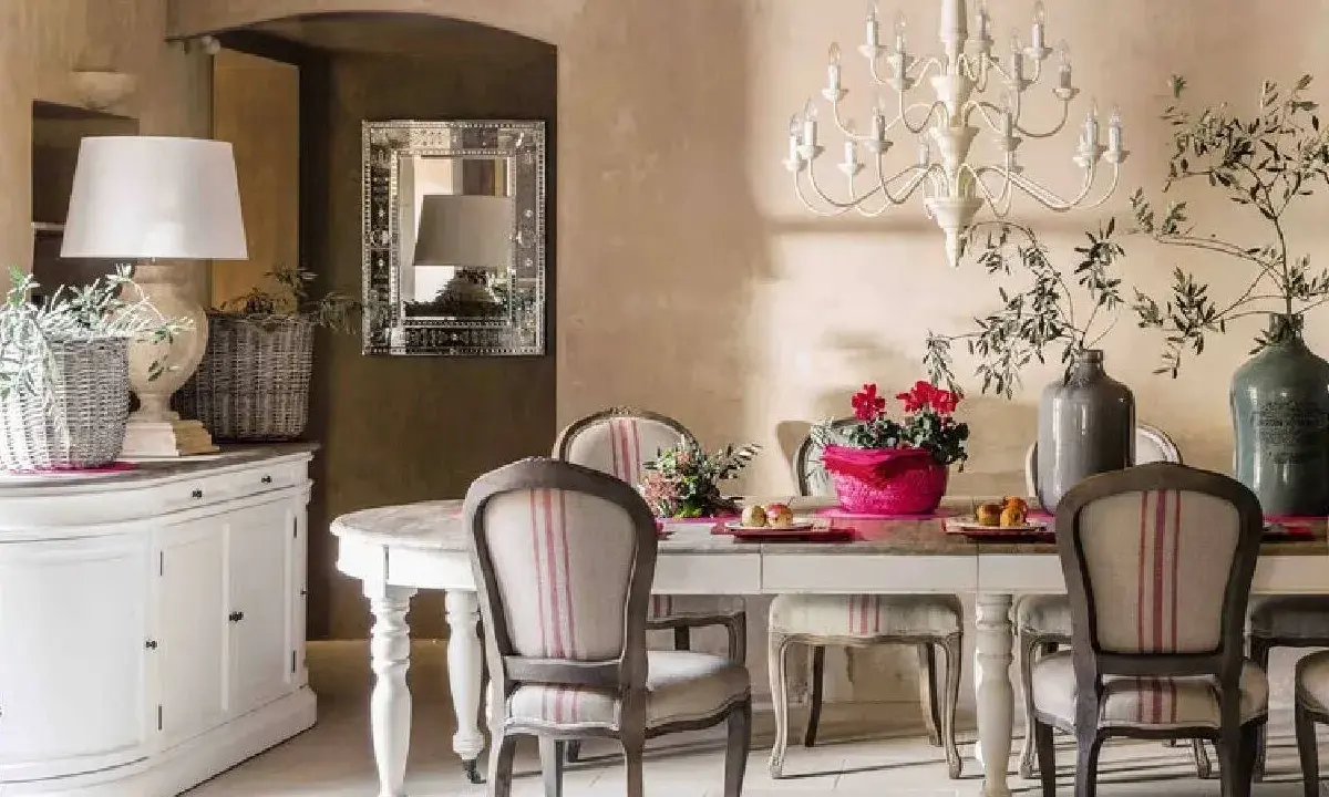 Comedor con estilo provenzal moderno. Mesa con sillas tapizadas, lámpara de araña, jarrón con ramas y flores. Aparador blanco a la izquierda.