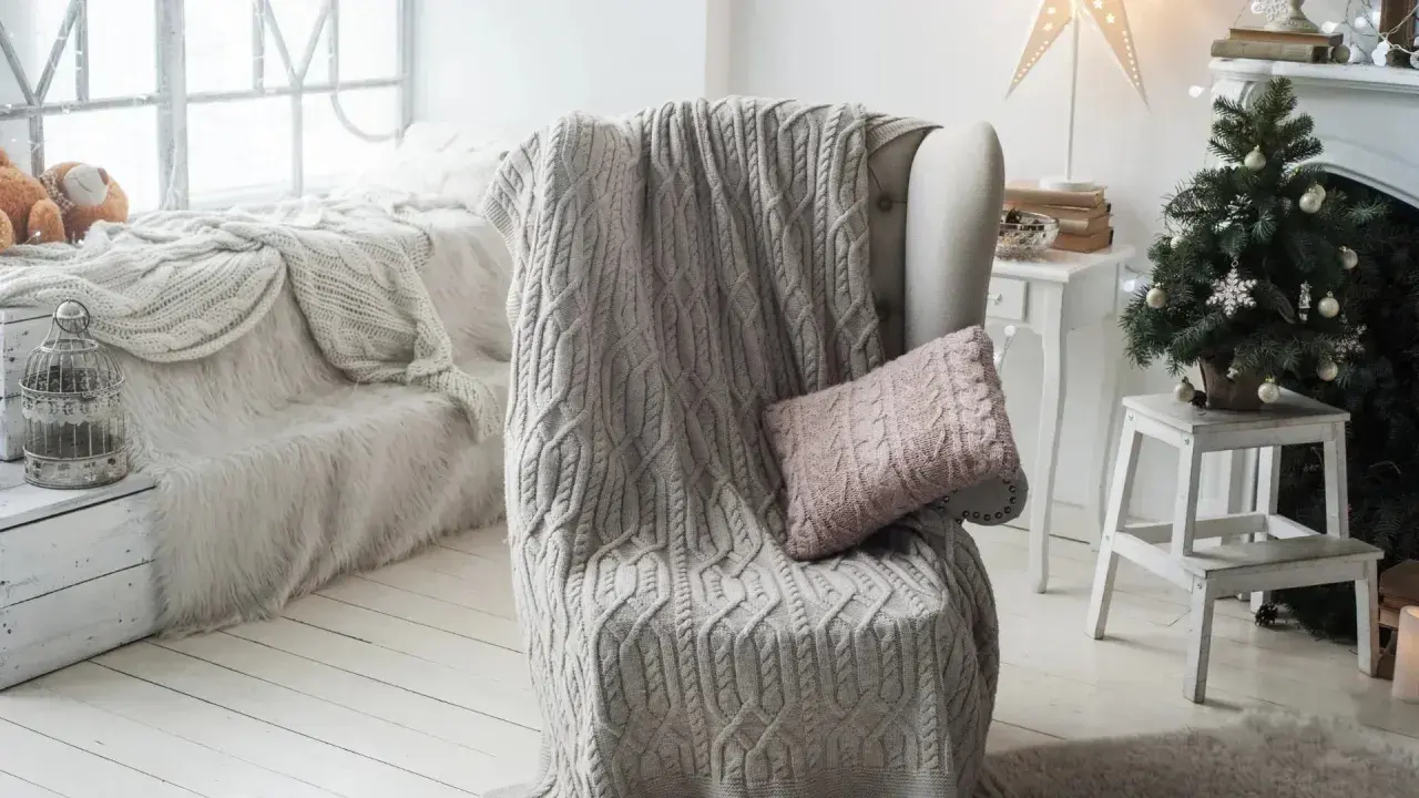 Manta de punto gris con trenzado, estilo plaid Bassetti, sobre sillón junto a árbol de Navidad.
