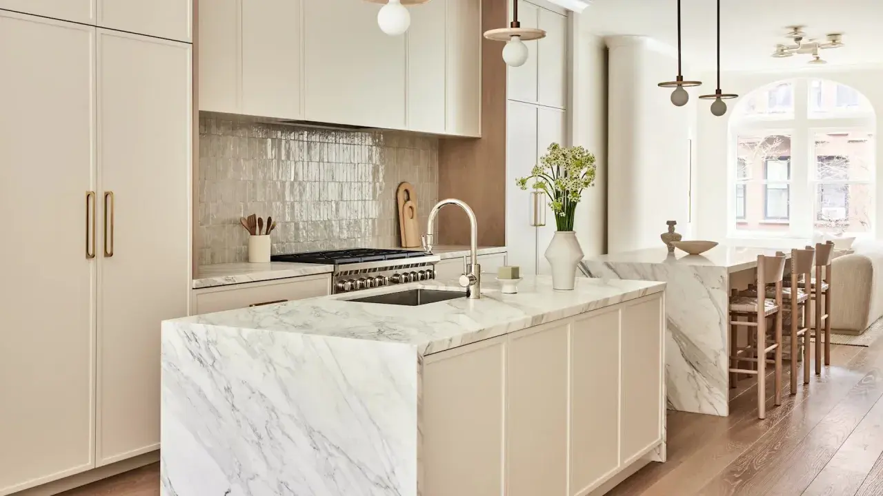 Elegante cocina atemporal con isla de mármol, gabinetes crema y electrodomésticos de acero inoxidable. Un jarrón con flores ilumina el espacio.