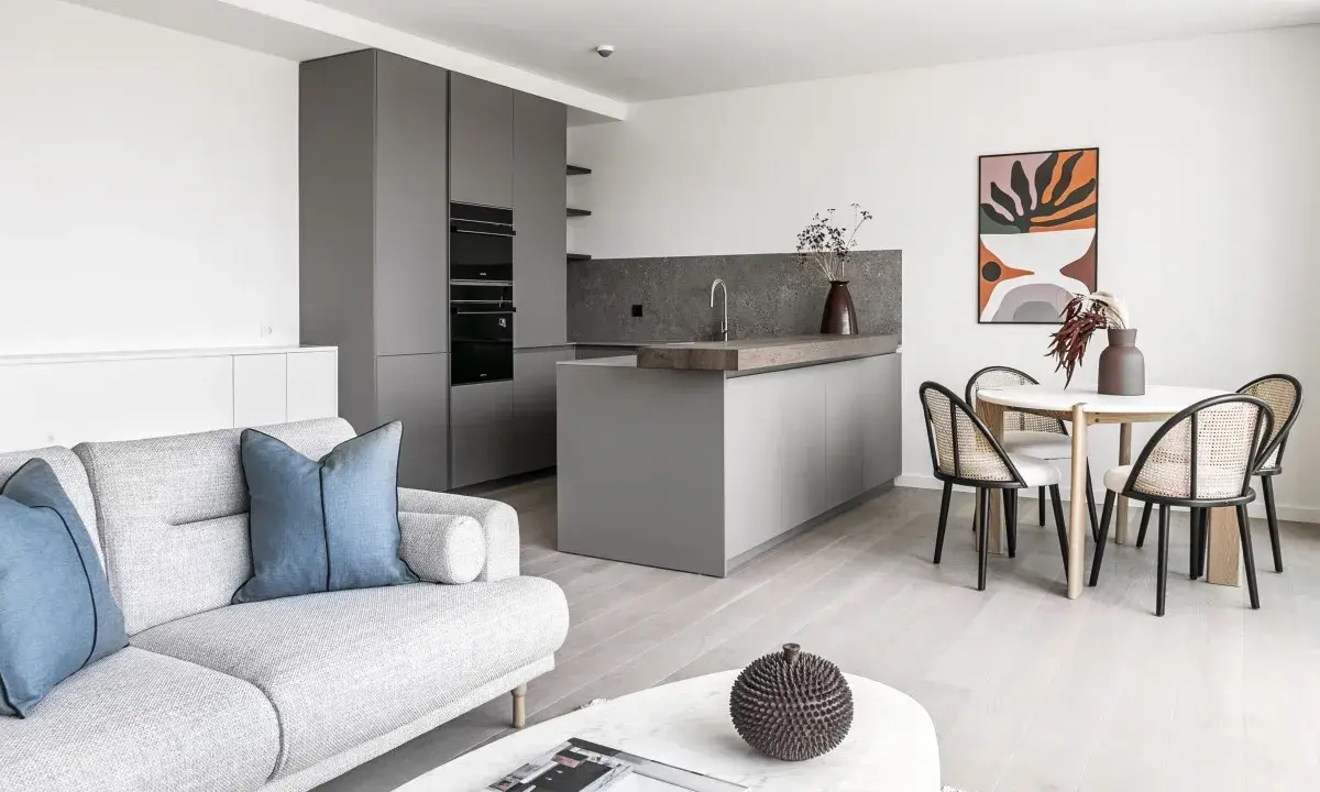 Salón moderno y cocina integrada, ideal para amueblar piso de alquiler. Sofá gris, mesa redonda con sillas de ratán y arte abstracto.
