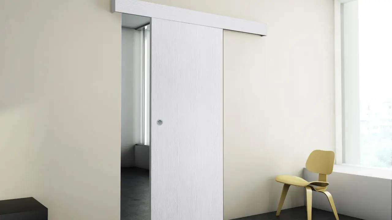 Puerta corredera blanca moderna instalada en pared beige, con silla amarilla y ventana al fondo.
