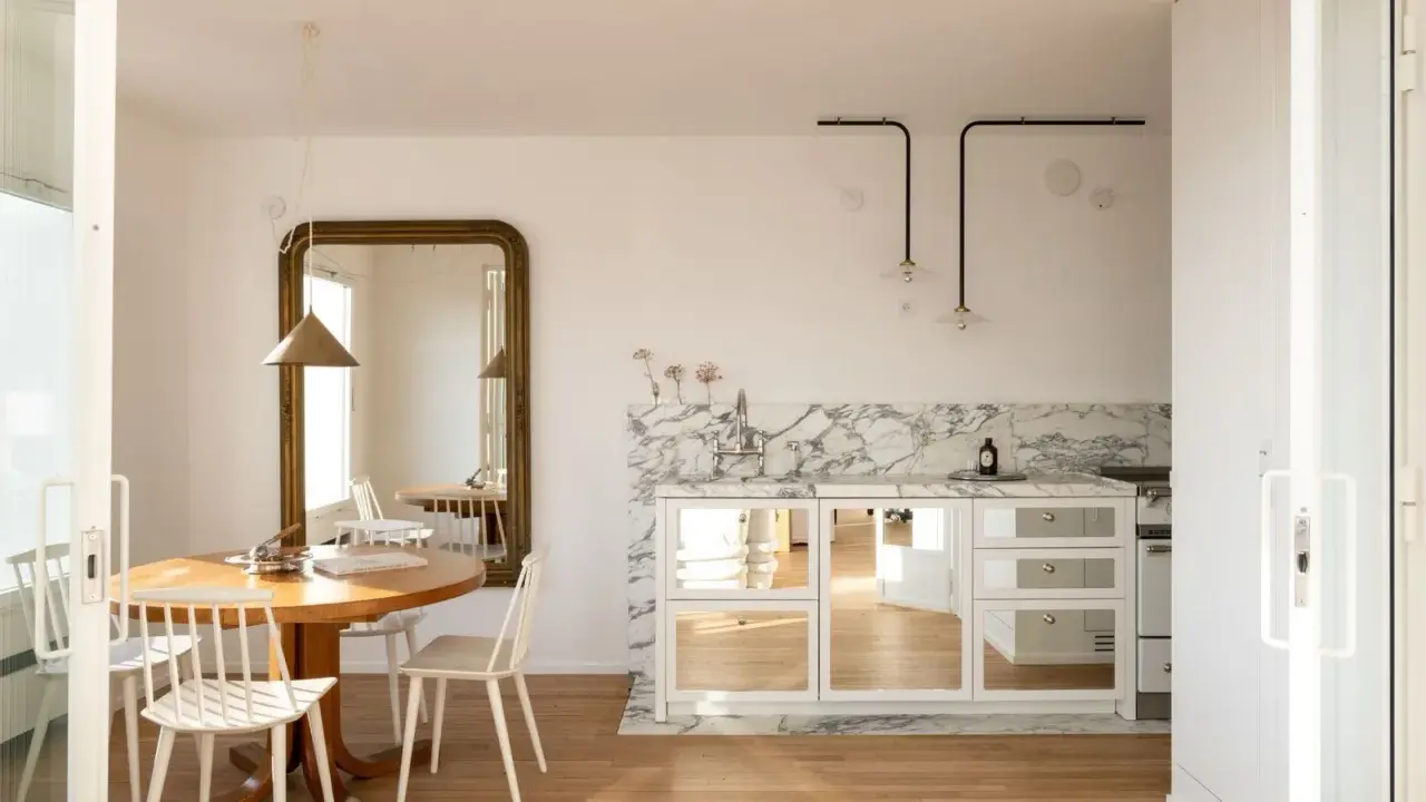 Comedores pequeños con encanto, con mesa redonda de madera, sillas blancas y un gran espejo dorado. La cocina integrada tiene encimera de mármol y armarios con puertas de espejo.