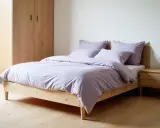 Cama con funda nórdica Bassols en tono lavanda, almohadas a juego, mesita de noche de madera y armario.