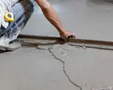 Obrero con botas de goma y cinta métrica en el bolsillo, usando una regla para nivelar suelo de cemento fresco.