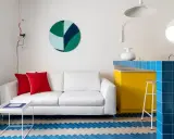Sofá blanco con cojines rojos, mesa auxiliar blanca y suelo con diseño de espiga en colores claros y azul. Barra de cocina amarilla y azul.