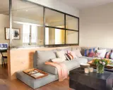 Un salón moderno con una pared de cristal que separa un área de trabajo. Un sofá gris con cojines coloridos y una manta rosa.