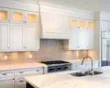 Cocina moderna con campana extractora decorativa sobre estufa. Gabinetes blancos, encimera de mármol y refrigerador de acero inoxidable.