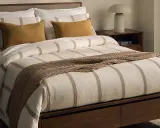 Cama con ropa de cama a rayas, cojines mostaza y manta tejida. Las bassols sabanas aportan un toque acogedor.