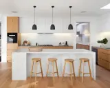 Cocinas modernas con isla blanca, taburetes de madera y gabinetes de roble. Escalera de diseño minimalista al fondo.