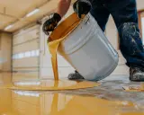 Persona con guantes vierte pintura amarilla de un cubo para pintar suelo cemento sin pulir.