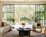 Salones elegantes con sofás curvos, una silla acogedora y grandes ventanales que conectan con un jardín de olivos.