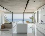 Moderna cocina americana y salón con vistas al mar. Isla blanca, sofá gris y suelo de mármol brillante.