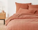 Cama con funda nordica terracota a cuadros y rayas. Mesita de noche de madera con lámpara y planta.