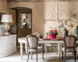 Comedor con estilo provenzal moderno. Mesa con sillas tapizadas, lámpara de araña, jarrón con ramas y flores. Aparador blanco a la izquierda.