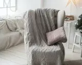 Manta de punto gris con trenzado, estilo plaid Bassetti, sobre sillón junto a árbol de Navidad.