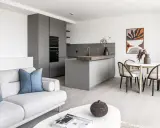 Salón moderno y cocina integrada, ideal para amueblar piso de alquiler. Sofá gris, mesa redonda con sillas de ratán y arte abstracto.