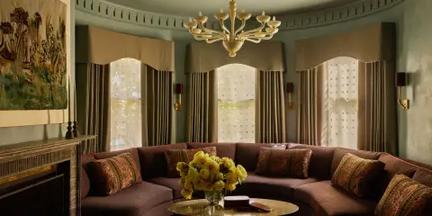 Sofá curvo marrón con cojines decorativos, mesa redonda con flores amarillas y un tapiz de estilo art déco en la pared.