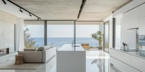 Moderna cocina americana y salón con vistas al mar. Isla blanca, sofá gris y suelo de mármol brillante.