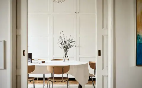 Comedor con mesa cubierta, sillas de madera y una pared en blanco RAL 9010. Un jarrón con ramas decora la mesa.