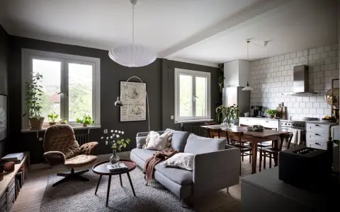 Un salón rectangular amueblado con sofá gris, sillón marrón, mesa de centro y alfombra. Cocina abierta con mesa de comedor.