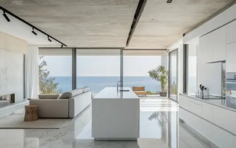 Moderna cocina americana y salón con vistas al mar. Isla blanca, sofá gris y suelo de mármol brillante.