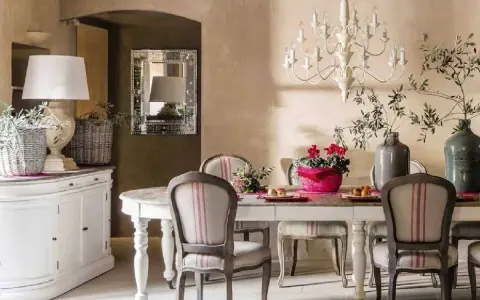 Comedor con estilo provenzal moderno. Mesa con sillas tapizadas, lámpara de araña, jarrón con ramas y flores. Aparador blanco a la izquierda.
