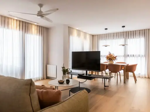 Acogedor salón de 20 metros cuadrados con sofá, mesa de centro, TV, y comedor. Cortinas blancas y suelo de madera crean un ambiente luminoso.