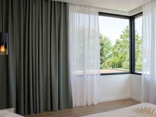 Cortinas bandalux grises y blancas en un dormitorio moderno, con vistas a un jardín frondoso.
