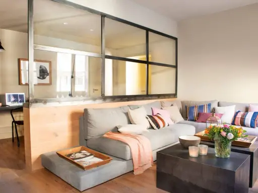Un salón moderno con una pared de cristal que separa un área de trabajo. Un sofá gris con cojines coloridos y una manta rosa.