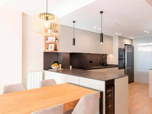 Moderna cocina en L con isla, electrodomésticos integrados, encimera de cuarzo y mesa de madera.
