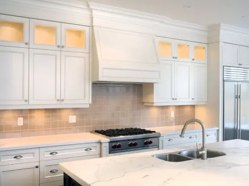 Cocina moderna con campana extractora decorativa sobre estufa. Gabinetes blancos, encimera de mármol y refrigerador de acero inoxidable.