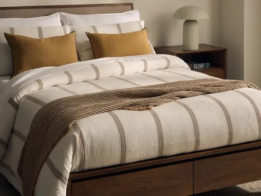 Cama con ropa de cama a rayas, cojines mostaza y manta tejida. Las bassols sabanas aportan un toque acogedor.