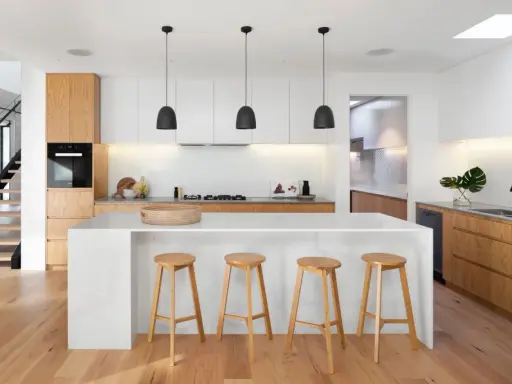 Cocinas modernas con isla blanca, taburetes de madera y gabinetes de roble. Escalera de diseño minimalista al fondo.