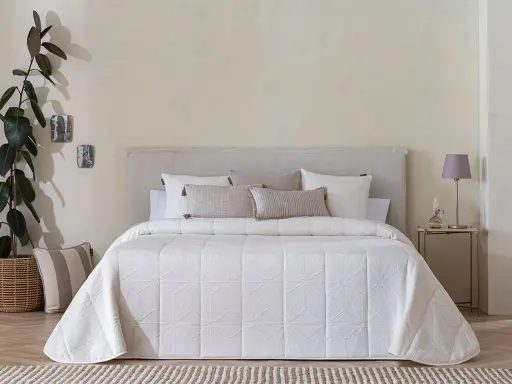 Una cama vestida con una colcha bouti beige de diseño geométrico, cojines y una planta decorativa.