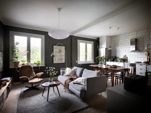 Un salón rectangular amueblado con sofá gris, sillón marrón, mesa de centro y alfombra. Cocina abierta con mesa de comedor.