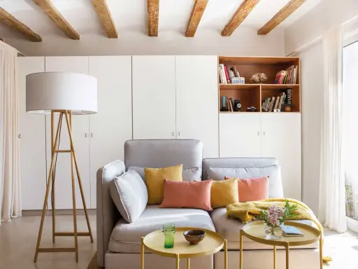 Ideas para decorar piso alquiler low cost: sofá gris, cojines coloridos, mesa auxiliar dorada, lámpara de pie y estantería con libros.