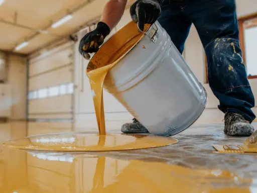 Persona con guantes vierte pintura amarilla de un cubo para pintar suelo cemento sin pulir.