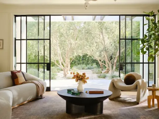 Salones elegantes con sofás curvos, una silla acogedora y grandes ventanales que conectan con un jardín de olivos.