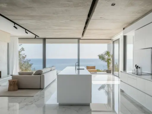 Moderna cocina americana y salón con vistas al mar. Isla blanca, sofá gris y suelo de mármol brillante.