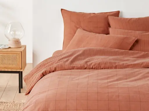 Cama con funda nordica terracota a cuadros y rayas. Mesita de noche de madera con lámpara y planta.